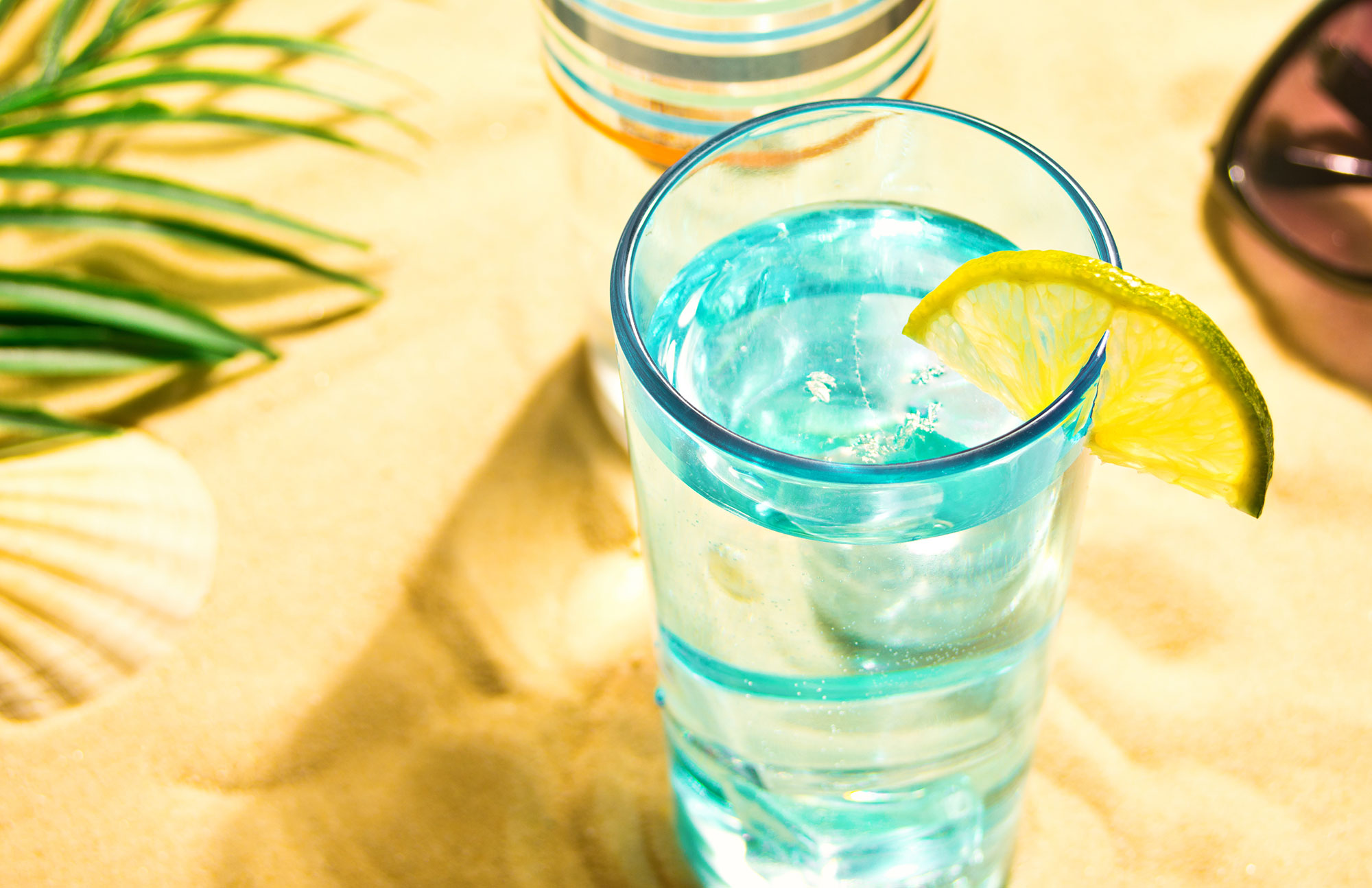 Glas Wasser mit Zitronenscheibe im Sand – erfrischendes Getränk gegen Sommerhitze